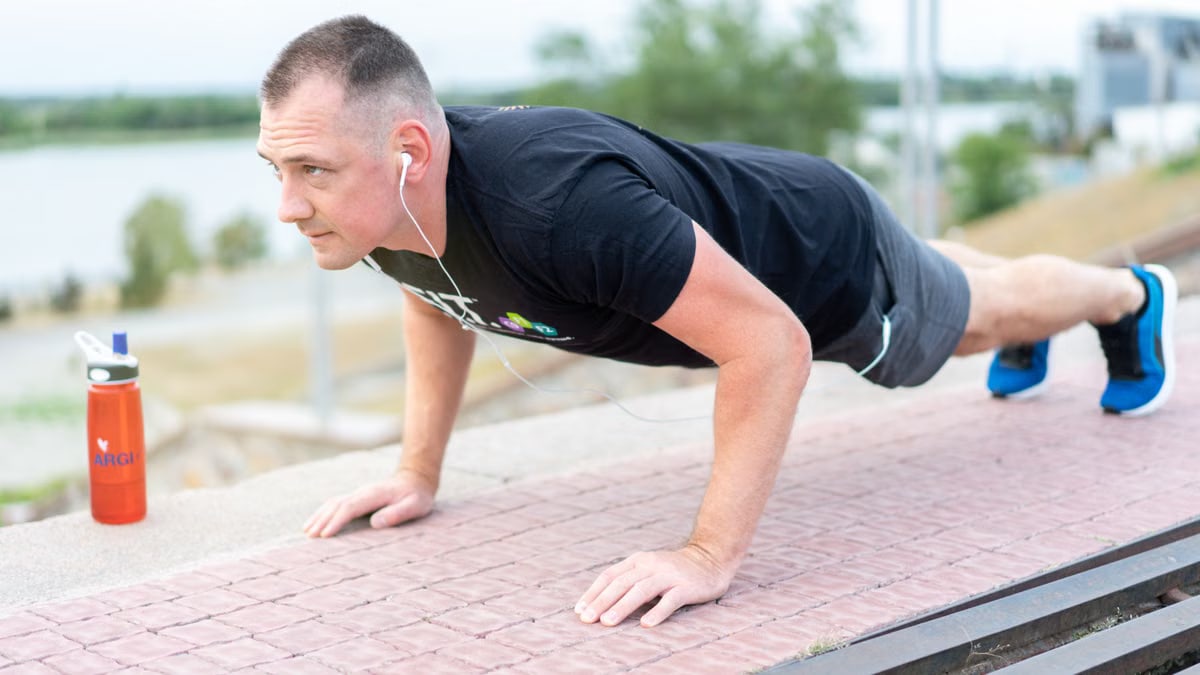 Adult male exercising outdoors performing push-ups with earphones and water bottle.